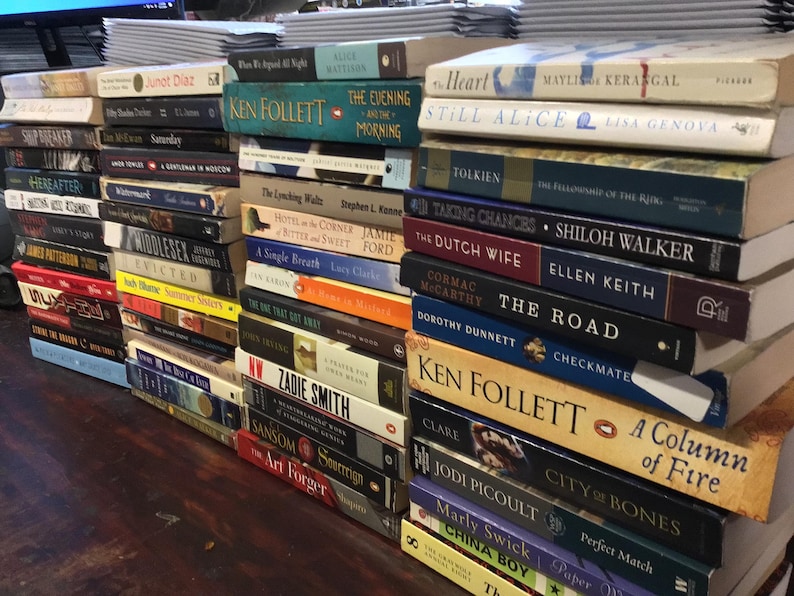 Op de afbeelding: Een stapel paperbacks met verschillende titels en auteurs. De boeken zijn verticaal gestapeld, met de titels naar buiten gericht. Enkele van de titels zijn "The Evening and the Morning", "Still Alice", "The Fellowship of the Ring", "The Road", "Checkmate", "A Column of Fire", "City of Bones" en "China Boy".