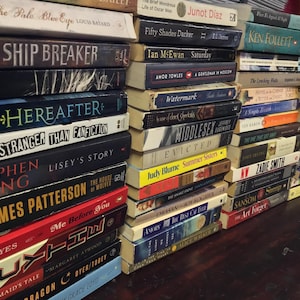 May include: A stack of paperback books with various titles, including "Ship Breaker", "Here After", "Stranger Than Fanfiction", "Lisey's Story", "The House of Wolves", "Me Before You", "The Handmaid's Tale", "Dragon Cryer/Taley", and "Grace Lloyd".