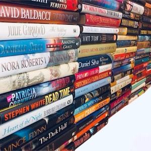 May include: A stack of paperback books with various titles and authors. The books are arranged in a staggered pattern, with the top of the stack leaning to the right. The books are all different colors and have different designs on their covers.
