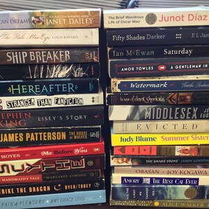 Op de afbeelding: Een stapel paperbacks met verschillende titels en auteurs. De boeken zijn verticaal gerangschikt, met de ruggen naar buiten. De titels omvatten "American Dreams", "Ship Breaker", "Here After", "Stranger Than Fanfiction", "Lisey's Story", "The House of Wolves", "Me Before You", "The Handmaid's Tale", "Strike the Dragon", "The Pain & Pleasure", "The Brief Wondrous Life of Oscar Wao", "Fifty Shades Darker", "Saturday", "A Gentleman in Moscow", "Watermark", "Middlesex", "Evicted", "Summer Sisters", "The Snake Stone", "Obasan", "The Best Cat Ever", "All the Light We Cannot See" en "The Color Purple".
