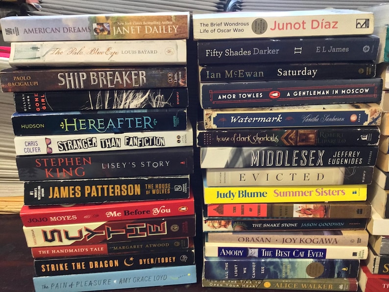 Op de afbeelding: Een stapel paperbacks met verschillende titels en auteurs. De boeken zijn verticaal gerangschikt, met de ruggen naar buiten. De titels omvatten "American Dreams", "Ship Breaker", "Here After", "Stranger Than Fanfiction", "Lisey's Story", "The House of Wolves", "Me Before You", "The Handmaid's Tale", "Strike the Dragon", "The Pain & Pleasure", "The Brief Wondrous Life of Oscar Wao", "Fifty Shades Darker", "Saturday", "A Gentleman in Moscow", "Watermark", "Middlesex", "Evicted", "Summer Sisters", "The Snake Stone", "Obasan", "The Best Cat Ever", "All the Light We Cannot See" en "The Color Purple".