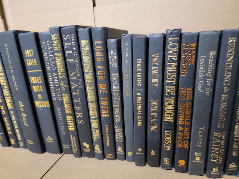 Pu&ograve; includere: Una collezione di libri con copertina rigida blu scuro e scritte dorate. I titoli includono "Love Must Be Tough", "Self Matters" e "Look for Me There". I libri sono disposti in fila, suggerendo una biblioteca o una libreria.