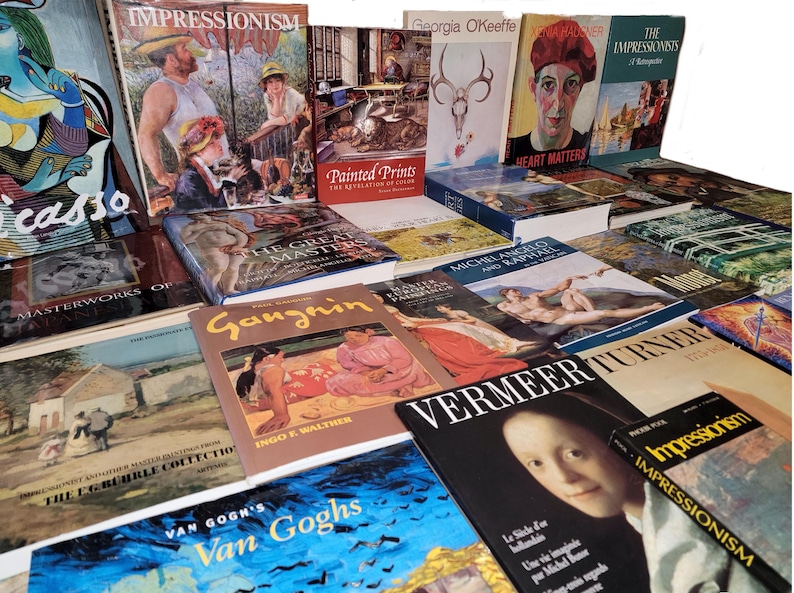 May include: A stack of art books featuring various artists, including Picasso, Impressionists, Georgia O'Keeffe, and Vermeer. The books are arranged in a staggered pattern, with the titles and authors visible.