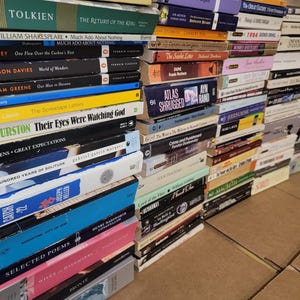 May include: A large stack of paperback books, mostly in shades of white, black, and brown, with some colorful titles. The books are arranged in a vertical stack, with the titles facing outward. The books are all different sizes and shapes, and some have different colored spines. The books are all in English.