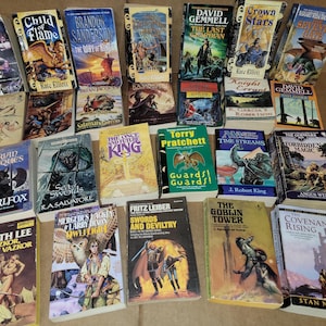 May include: A collection of fantasy and science fiction paperback books with various titles and authors. The books are stacked in a pile on a brown surface.