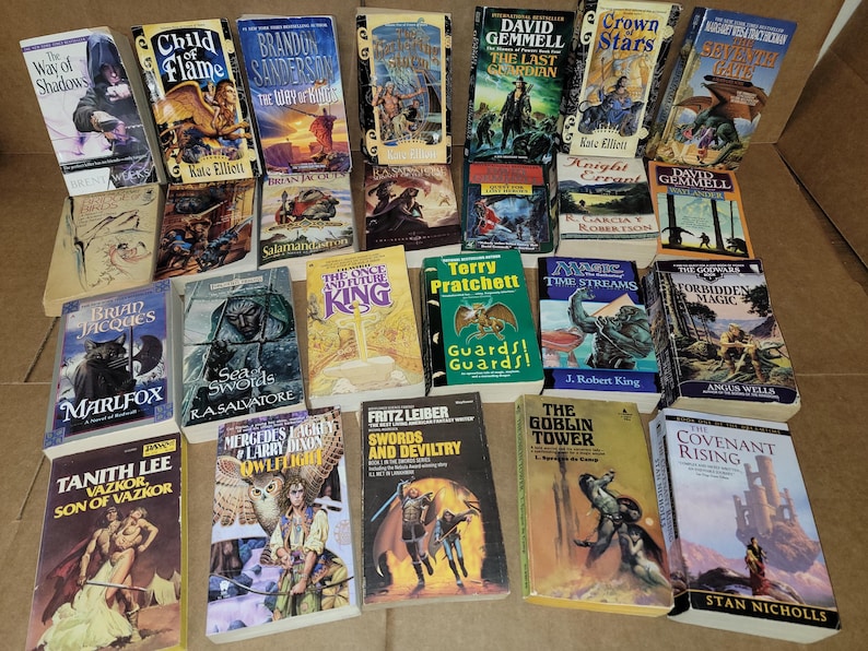 May include: A collection of fantasy and science fiction paperback books with various titles and authors. The books are stacked in a pile on a brown surface.