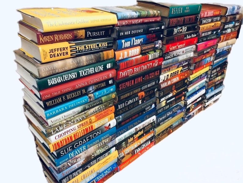 May include: A large stack of paperback books, mostly fiction, with various authors and titles visible. The books are arranged in a vertical stack, with the spines facing outward. The stack is on a white surface.