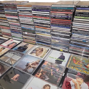 May include: A large collection of CDs, stacked in multiple piles. The CD cases display various album covers with images of musicians and album titles. The CDs are in clear plastic cases, and the spines are visible.
