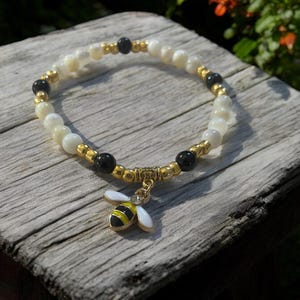 May include: A white and black beaded bracelet with a gold bee charm. The bracelet is made with small white and black beads and has a gold clasp. The bee charm is gold with black and yellow stripes.