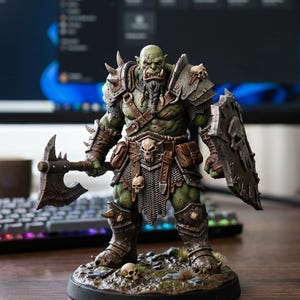 May include: A green-skinned orc warrior figurine standing on a textured base with skulls. The figure wears brown leather and metal armor, carries a large battle axe in the right hand, and holds a shield with a skull emblem in the left hand.