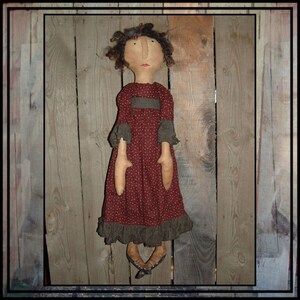 May include: A handmade rag doll with brown hair, a red dress with white polka dots, and black shoes. The doll is standing in front of a wooden background.