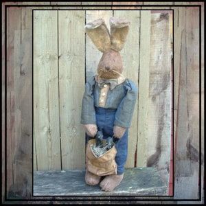 Puede incluir: Un conejo marrón con un traje azul y gris y una pajarita marrón sostiene una cesta de conejos marrones más pequeños. El conejo está de pie sobre una superficie de madera frente a una valla de madera.