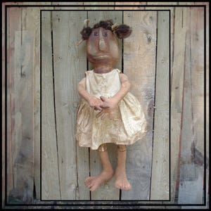 May include: A handmade rag doll with brown skin, brown hair, and a white dress. The doll is standing in front of a wooden fence.
