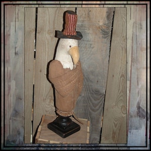 May include: A handmade patriotic eagle figurine with a red, white, and blue top hat. The eagle is made of fabric and has a brown body with a white head. The eagle is standing on a black base.