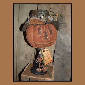 May include: A brown fabric pumpkin with a stitched-on face and a black hat sits on a black pedestal with a brown tag that says "Happy". The pumpkin is decorated with dried leaves and twigs.