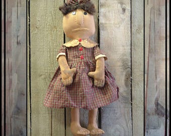 May include: A handmade rag doll with brown hair, a plaid dress, and red buttons. The doll has a simple, sweet expression and is standing against a wooden background.