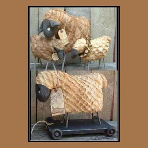 May include: Handmade sheep figurines with textured bodies and black faces. One sheep wears a floral crown, while another is on a wheeled cart. Each sheep has a tag attached. The sheep are displayed against a wooden background.