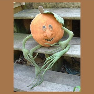 May include: A handmade orange pumpkin with a friendly face and long green arms sits on wooden steps. The pumpkin has a stitched smile and two black eyes.