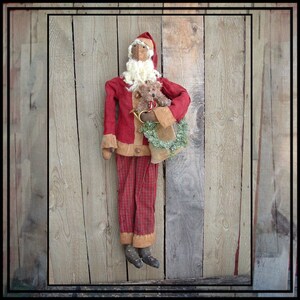 May include: A handmade Santa Claus doll with a red suit, plaid pants, and a white beard. The doll holds a small teddy bear and a green wreath. The Santa doll has a tan face and is wearing black boots. The background is a wooden wall.
