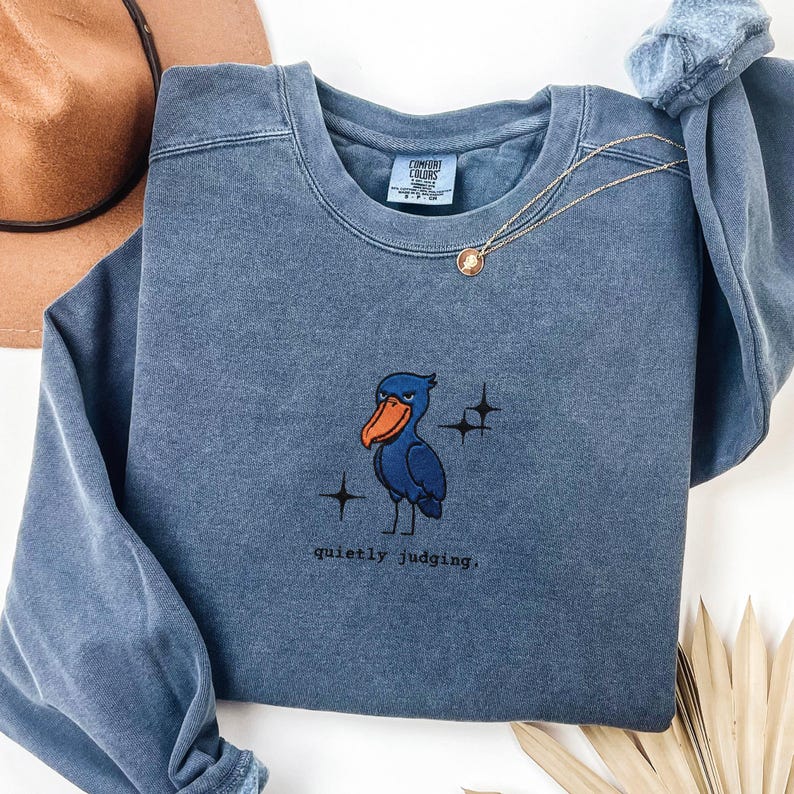 Op de afbeelding: Een blauwe Comfort Colors sweatshirt met een cartoon pelikaan ontwerp en de tekst "quietly judging". De pelikaan heeft een blauw lichaam en een feloranje snavel. Een gouden ketting is over de sweatshirt gedrapeerd.