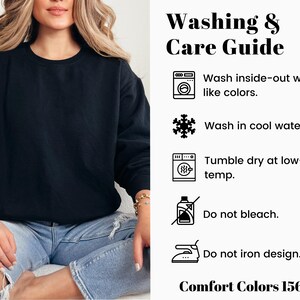 Op de afbeelding: Een zwarte crewneck sweatshirt met een was- en verzorgingsgids. De gids bevat instructies om binnenstebuiten te wassen met dezelfde kleuren, te wassen in koud water, te drogen in de droger op lage temperatuur, niet te bleken en het ontwerp niet te strijken. De tekst "Comfort Colors 1566" is ook zichtbaar.