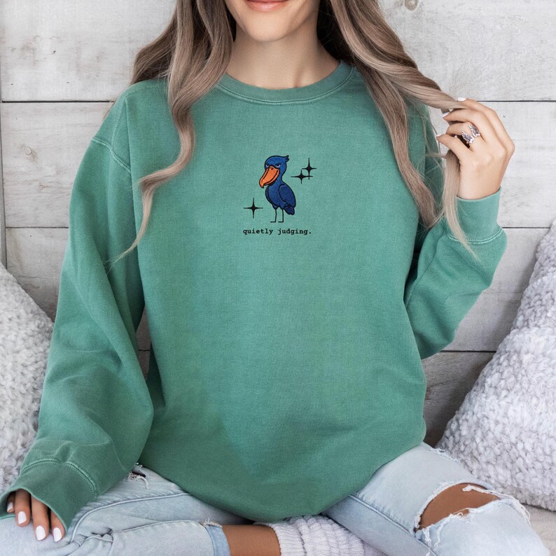 Op de afbeelding: Een tealkleurige sweatshirt met een cartoon vogelontwerp in blauw en oranje, met de tekst "quietly judging". De vogel wordt omringd door twee sterren graphics. De sweatshirt heeft lange mouwen en een ronde hals.