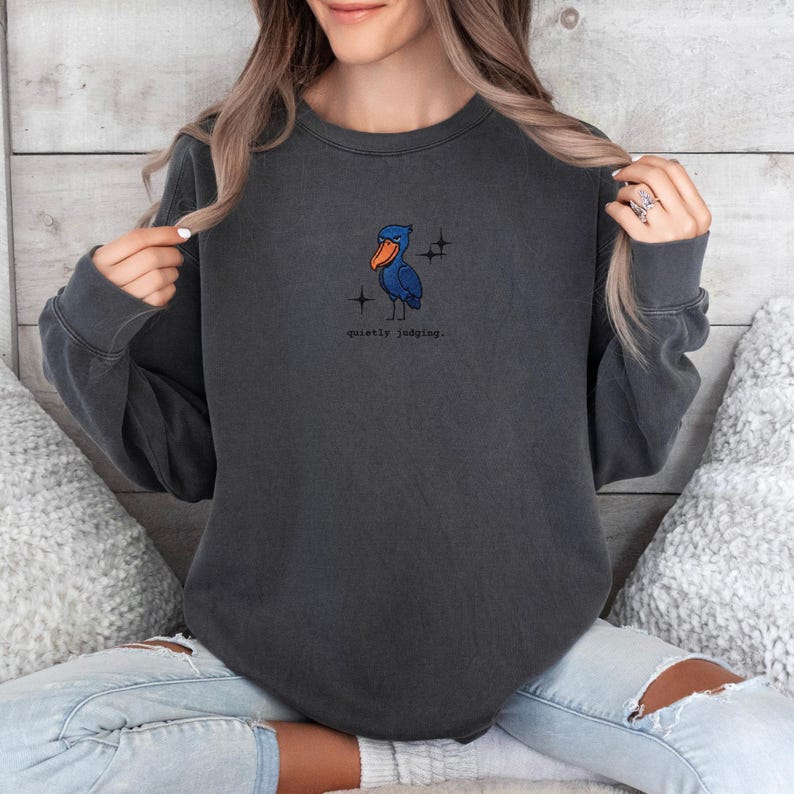 Op de afbeelding: Donkergrijze sweatshirt met een cartoon illustratie van een blauwe vogel met een oranje snavel en de tekst "quietly judging." De vogel is omringd door twee kleine sterren. De sweatshirt heeft lange mouwen en een ronde hals.