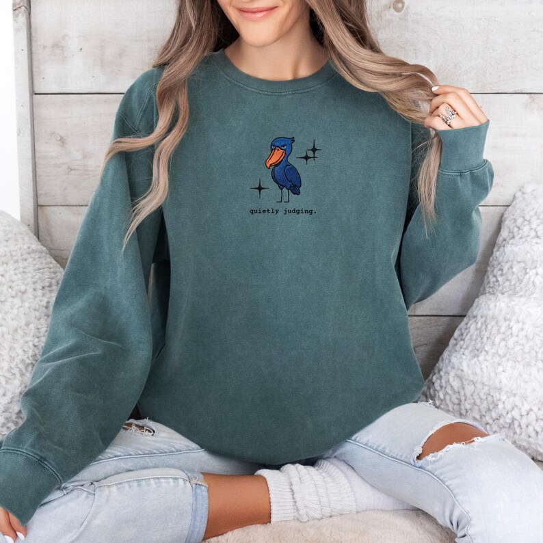 Op de afbeelding: Tealkleurige sweatshirt met een klein geborduurd pelikaanontwerp en de tekst "quietly judging". De pelikaan is blauw met een oranje snavel en het ontwerp bevat twee kleine stervormige vormen.