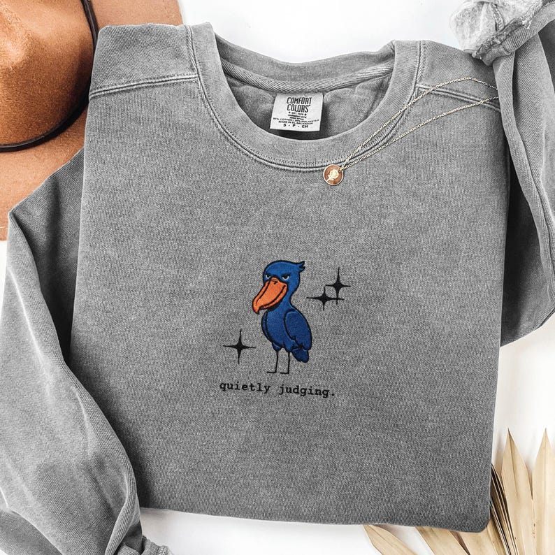Op de afbeelding: Een grijze Comfort Colors sweatshirt met een geborduurde blauwe vogel met een oranje snavel en de tekst "quietly judging". Het ontwerp bevat twee stervormige graphics. Een gouden ketting is zichtbaar.
