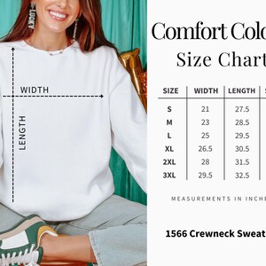 Op de afbeelding: Witte crewneck sweatshirt met een maattabel. De tabel geeft de afmetingen in inches voor breedte, lengte en mouwlengte, vari&euml;rend van maat small tot 3XL. De tekst "Comfort Colors" en "1566 Crewneck Sweatshirt" zijn ook zichtbaar.