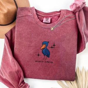 Op de afbeelding: Een bordeauxrood Comfort Colors sweatshirt met een geborduurde blauwe vogel, een oranje snavel en de tekst "quietly judging". Een gouden ketting met een hanger ligt op het sweatshirt. Het sweatshirt is op een wit oppervlak gevouwen.