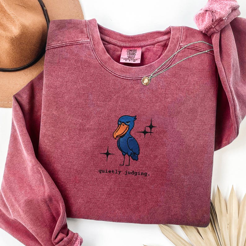 Op de afbeelding: Een bordeauxrood Comfort Colors sweatshirt met een geborduurde blauwe vogel, een oranje snavel en de tekst "quietly judging". Een gouden ketting met een hanger ligt op het sweatshirt. Het sweatshirt is op een wit oppervlak gevouwen.