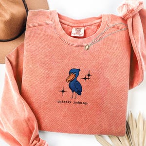 Op de afbeelding: Koraalrood sweatshirt met een ronde hals en lange mouwen. De voorkant heeft een geborduurde blauwe vogel met een oranje snavel, sterren en de tekst "quietly judging."