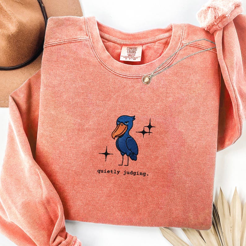 Op de afbeelding: Koraalrood sweatshirt met een ronde hals en lange mouwen. De voorkant heeft een geborduurde blauwe vogel met een oranje snavel, sterren en de tekst "quietly judging."