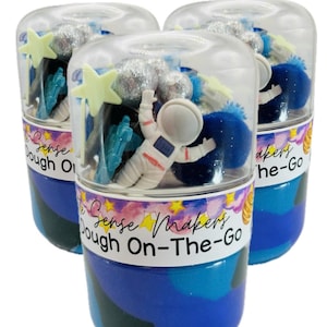 May include: Three clear plastic containers filled with space-themed sensory dough. Each container has an astronaut figurine, star and rocket-shaped toys, and the text "Sense Makers Dough On-The-Go". The containers have blue and silver accents.