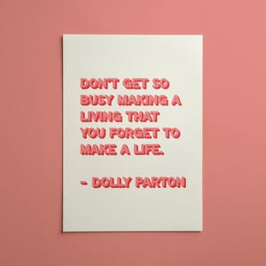 May include: A rectangular white greeting card featuring the red text: DON'T GET SO BUSY MAKING A LIVING THAT YOU FORGET TO MAKE A LIFE. - DOLLY PARTON. The lettering has a slight 3D effect with a darker red shadow on the right side of each letter.