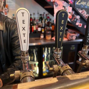 Puede incluir: Dos grifos de cerveza con la palabra "TEXTE" escrita en ellos. El grifo de la izquierda es blanco con letras negras y el grifo de la derecha es negro con letras doradas.