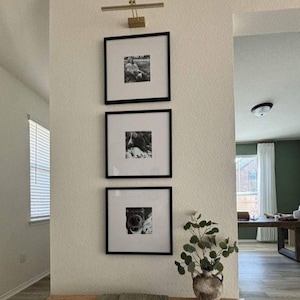 Pode incluir: Três molduras quadradas pretas com paspatur branco estão dispostas verticalmente numa parede, cada uma contendo uma fotografia a preto e branco. Uma luz de quadro em tom dourado está montada acima da moldura superior, iluminando a arte.
