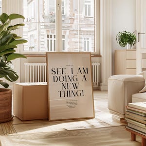May include: A framed print with the text "SEE, I AM DOING A NEW THING!" in black lettering. The print is propped up against a light brown box on a wooden floor. A large green plant and a window are in the background.