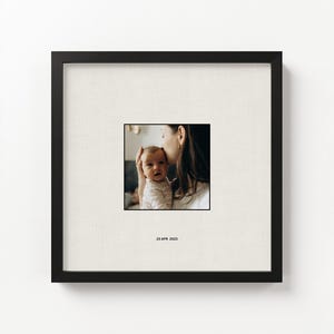 Op de afbeelding: Een vierkante zwarte fotolijst met een gecentreerde foto van een persoon die een baby op het voorhoofd kust. De foto is geplaatst op een getextureerd gebroken wit passe-partout met de tekst 23 APR 2023 onder de afbeelding.