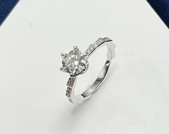 May include: A silver ring with a large, round, clear gemstone surrounded by smaller clear gemstones. The ring is on a white hexagonal display stand.