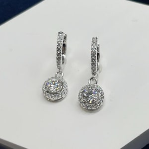 Könnte beinhalten: Ein Paar silberne Creolen mit einem baumelnden runden, diamantförmigen Stein, der von einem Heiligenschein aus kleineren Diamanten umgeben ist.