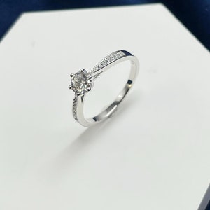 May include: A silver ring with a large, round, clear gemstone as the focal point. The band is thin and features small, clear accent stones on the sides. The ring is displayed on a white, hexagonal surface against a dark blue backdrop.