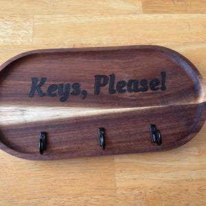 May include: A wooden key holder with the text "Keys, Please!" engraved on it. The holder has three hooks for hanging keys.