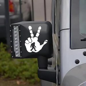 May include: A black side view mirror with a white sticker of a Bigfoot hand giving the peace sign. The sticker is on a Jeep.