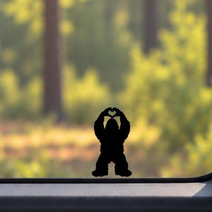 Peut inclure: Un autocollant en vinyle noir représentant la silhouette d'une créature Bigfoot debout. La créature a les bras levés au-dessus de la tête, ses mains étant positionnées pour former une forme de cœur. L'autocollant est appliqué sur une vitre.