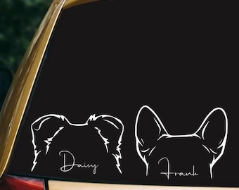 May include: A white vinyl decal applied to a car window featuring the line-art silhouettes of two dog heads. The left silhouette has floppy ears with the name Daisy written in script, and the right silhouette has pointed ears with the name Frank.