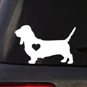 May include: A white Basset Hound dog decal with a heart detail, applied to a car window. The dog is shown in profile. The decal is a simple silhouette, suitable for vehicle decoration.