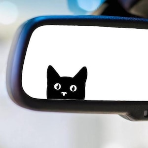 May include: A black cat sticker peeking over the edge of a car's rearview mirror. The cat has a simple, cartoon design with large eyes and a small nose. The mirror is black with a white reflective surface.