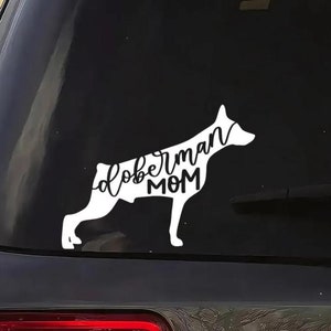 May include: A white vinyl decal of a Doberman silhouette with the words "Doberman MOM" in a cursive font. The decal is affixed to the rear window of a vehicle, showcasing a dog-themed design.